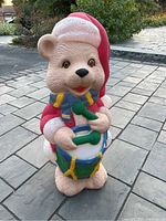 Front view of vintage teddy bear blow mold in Santa outfit holding drumsticks and standing on paved surface outdoors.