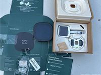 Ecobee thermostat parts including the white mounting base, external room sensor, screws, installation manual, and original box packaging display.
