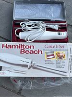 Open carrying case showing electric knife handle, power cord coiled, two cutting blades and carving fork, all organized in a molded red tray inside the silver case lid lined with bubble wrap.