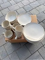 Overall view of the set showing stacked dinner plates, bowls, and mugs on a cardboard box outside on a tile floor.
