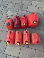 Eight red used plastic gas cans with various black and yellow caps arranged on stone pavement. All cans have signs of use and dirt. Different sizes and shapes visible including square and rectangular cans.