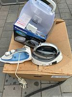 Two electric irons shown on a box; one Panasonic in white and black with clear water tank, one Sunbeam in white and blue with cord coiled.