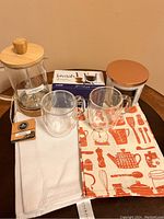 Overview of lot showing glass tea maker with wooden lid, two double wall coffee cups, decorative tea towel, white towel, and metal candle tin with copper lid.