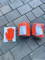 Two sealed bundles of bright orange gloves with one glove laid out showing criss-cross pattern and knit construction.