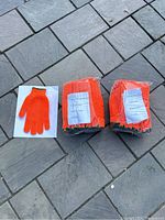 Two sealed bundles of bright orange gloves with PVC coating, accompanied by a single glove displayed on white paper for detail showing the knit and coating pattern.