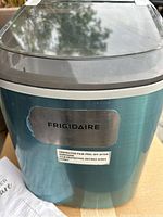 Front view showing Frigidaire logo and protective film label on metallic blue portable ice maker