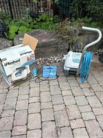 Full view of the lot showing the pool cleaning robot, its box, blue charger, and wheeled caddy with hose, set on paved area outdoors.