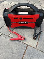Front angle showing the red and black battery booster with one red and one black clamp attached to red and black jumper cables resting on stone tile ground.