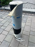 Photo of rolled roofing membrane wrapped in gray plastic showing the roll standing upright on a paved surface.