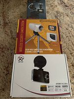 Photo showing the Centurion wireless security camera system box with camera mounted on tripod and two DM100 dash camera boxes placed behind.