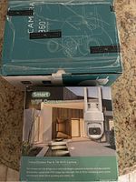 Two boxes of smart WiFi cameras, stacked. Boxes label the product as 'Smart WiFi Camera', showing images of the white pan and tilt camera with dual antennas. Includes branding details like 'Indoor/Outdoor Pan & Tilt Wi-Fi Camera' and mentions full HD crystal clear image day and night with smart alerts and 360-degree coverage.