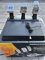 Front view of Thrustmaster T-LCM racing pedals with three pedals and attached cable, shown on top of the original box.