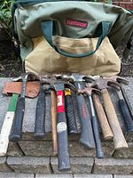 Photo showing 7 hammers, one axe with leather sheath, and the large Storehouse tool bag behind them.