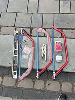 Photo showing three new bow saws with red frames and blue blades, a used metal level, locking vise grips, a bag of Bondex Plaster of Paris, new gloves, and ear plugs, all arranged on stone tile ground.