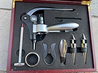 Photo of wooden box containing wine and liquor pouring set including corkscrew, thermometer, bottle stoppers, pour spout, foil cutter, and bottle clip with foam inserts.