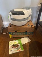 T-Fal Actifry white air fryer on a cardboard box with instruction manual visible below.