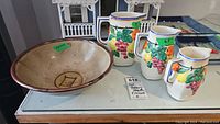 Photo showing signed pottery bowl and three colorful fruit decorated pitchers arranged on glass surface.