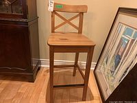 Wooden bar chair with cross-back and reinforced lower rung for feet support, placed on wooden floor next to furniture and a framed painting.