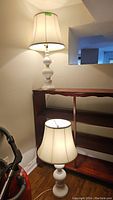Photo of two white ceramic table lamps with beige fabric shades, one standing on the floor and one placed on a wooden shelf.