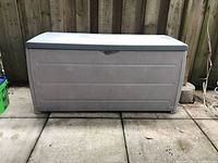 Front view of the Black & Decker resin deck box placed on concrete tiles against a wooden fence, showing light dirt and wear.