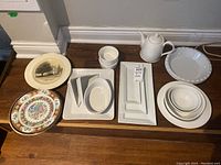 Overview of assorted ceramic dishes including decorative plates, white serving trays, ramekins, pie plates, and teapot arranged on wooden surface.