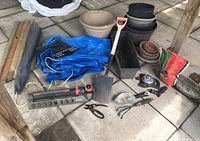 Wide view showing various garden tools, planters, terra cotta pot trays, tarp and other supplies placed on concrete floor under a wooden structure.