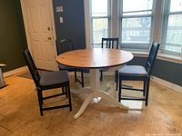 Round wood kitchen table with white pedestal base and natural wood top, surrounded by four black chairs with cushions. Tables and chairs show signs of wear and scuffs.