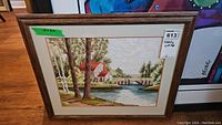 Framed needlework artwork showing a landscape with a river, bridge, trees, and red-roofed houses.