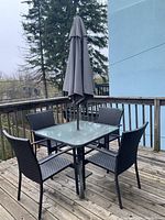 Full view of square glass top outdoor table with four woven upholstery chairs and umbrella on wooden deck