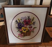 Framed pansy needlepoint artwork with colorful flowers in a basket, wood frame and glass cover, shown standing against a wooden floor.