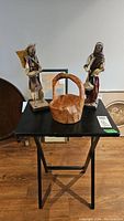 Two paper mache figurines standing on a black folding TV tray with a wood carved basket in front.