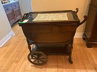 Side angle view of a wooden tea cart with glass top and decorative cloth inside top frame, showing one large wheel and folding side panels.