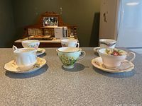 Overview of six porcelain teacups and saucers with floral patterns and gold trim arranged on countertop.
