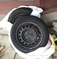 Two Bridgestone tires with metal coated rims stacked and wrapped in a white plastic bag on concrete surface.