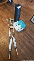 Photo showing two aluminum crutches, black yoga mat rolled up, and Dr. Scholl's foot bath on hardwood floor.