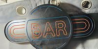Black circular wall sign with orange neon-style BAR text surrounded by side extensions with matching orange outline designs.