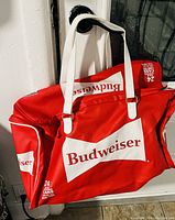 Red insulated Budweiser cooler bag with white handles and logos, shown hanging on a door knob.