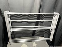 Foldable white clothes drying rack with metal rods and plastic connectors, shown set up in front of a curtain.