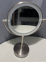 Photo shows a round double sided makeup mirror, metal frame and base, placed on a white surface against a dark background.