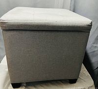Closed square grey fabric ottoman, showing tufted padded top and four black legs.