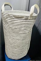 Photo of cylindrical white fabric hamper with black horizontal stripes and two white handles on sides.