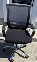 Front view of one black mesh office computer chair with padded seat, armrests, and chrome base with wheels on a concrete floor.
