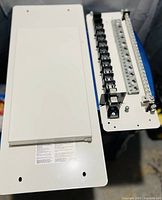 White metal fusible load center insert with multiple fuse holders and terminal strip, removable cover panel placed beside it.