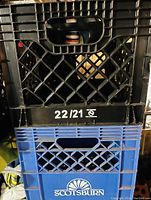 Photo showing two plastic milk crates stacked; top black crate with 22/21 marking, bottom blue crate with Scotsburn branding.