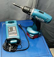 Photo shows blue Makita cordless drill with drill bit attached alongside a matching blue Makita charger with attached power cord, both on a blue surface with a dark backdrop.