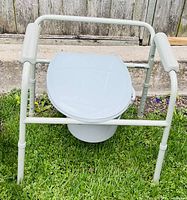 Front view of a mobility toilet seat showing white plastic seat and lid on an adjustable white metal frame with padded armrests. Bucket is visible underneath the seat.