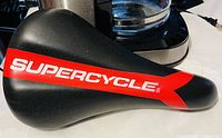 Black bicycle seat with red and white Supercycle branding on the side, photographed on a surface indicating new condition.
