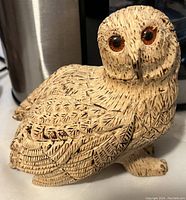 Photo of a small owl figurine with carved feather details and reddish-brown eyes on a flat surface with a metallic object behind.