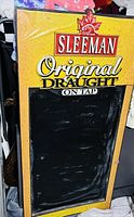 Single photo of a freestanding chalkboard advertising sign with Sleeman Original Draught branding on top in red, yellow, and black colors and a large blank blackboard section underneath.
