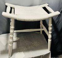 White vintage stool with woven seat, turned white wooden legs and crossbars, shown on plain surface.